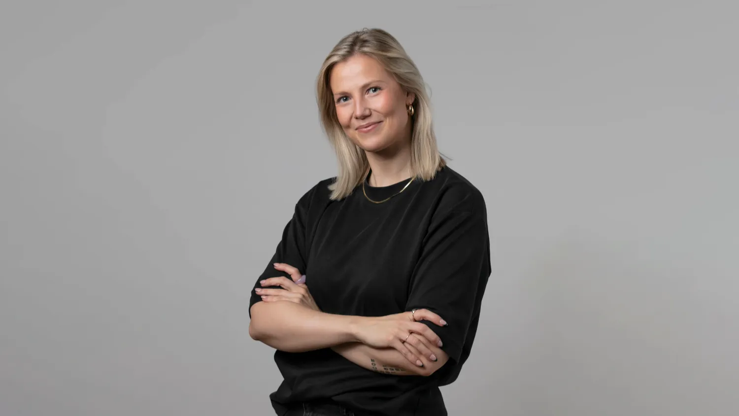Kuva Legit AI Oy:n toimitusjohtaja Netta Heikkilästä. Netalla on päällään tumma t-paita, tummat housut ja kädet ovat ristissä. Netta hymyilee kameralle. Tausta on neutraali.